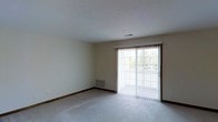 This single-floor space offers a clean, functional layout designed for comfortable everyday living. The main living area is finished with wall-to-wall carpeting in a neutral tone, contributing to a warm and understated aesthetic throughout. Smooth, light-colored walls provide a versatile backdrop, while dark wood baseboards add a subtle contrast and a touch of classic detailing.

A prominent sliding glass door serves as both a key architectural feature and a source of abundant natural light, drawing the eye and brightening the interior. Dressed with vertical blinds for adjustable privacy and light control, the door opens to an outdoor area, offering a seamless connection between the interior and exterior. Greenery is visible through the glass, adding a touch of nature to the view.

Ceiling-mounted HVAC vents ensure efficient climate control throughout the space, while a floor-level baseboard heating unit provides an additional layer of comfort. The ceiling is smooth and well-maintained, contributing to the overall clean and uncluttered feel of the space. This straightforward, move-in-ready layout presents an excellent opportunity for personalization and customization to suit a variety of lifestyles.