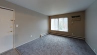 This single-floor unit offers a practical and comfortable layout across several well-defined spaces. The living area welcomes you upon entry through a secured front door, featuring neutral beige walls, plush gray carpeting, and a large picture window fitted with vertical blinds that allows generous natural light and views of the surrounding greenery. Baseboard heating units run along the walls throughout the unit, providing consistent warmth.

The galley-style kitchen is efficiently designed with ample white cabinetry lining both sides, light countertops, tile flooring, and black appliances including an electric range and a full-size refrigerator. A window with vertical blinds and an under-cabinet range hood complete the space, while a sink with a double basin sits along the opposite counter wall.

The bedrooms are carpeted and finished with neutral walls, each featuring windows with vertical blinds and baseboard heating. At least one bedroom includes a closet with bifold doors, offering built-in storage with a shelf and hanging rod. Tile flooring at the closet entrance provides a practical transition.

The bathroom features a full tub-and-shower combination with white tile surround and a frosted glass window above the tub, offering privacy while still allowing natural light. Overall, the unit presents a clean, functional living environment with consistent finishes throughout.