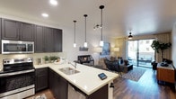 This single-floor residence offers a thoughtfully designed open-concept layout that seamlessly connects the kitchen and living areas. The kitchen is a standout feature, showcasing dark charcoal cabinetry paired with crisp white quartz countertops that extend into a peninsula with a double-basin sink. Three elegant pendant lights hang above the peninsula, adding both function and style, while recessed lighting illuminates the space evenly throughout. A classic subway tile backsplash adds a clean, modern touch to the kitchen's aesthetic.

The open layout flows naturally into the living area, which is anchored by warm wood-tone flooring that runs throughout the space. The living room is comfortably furnished and benefits from abundant natural light streaming in through large sliding glass doors that lead to an outdoor area, creating a bright and airy ambiance. A ceiling fan enhances comfort, while sheer curtains soften the natural light. The overall design strikes a balance between contemporary style and everyday functionality, with neutral tones, modern fixtures, and cohesive finishes creating a welcoming and well-appointed living environment.