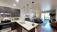This single-floor residence offers a thoughtfully designed open-concept layout that seamlessly connects the kitchen and living areas. The kitchen is a standout feature, showcasing dark charcoal cabinetry paired with crisp white quartz countertops that extend into a peninsula with a double-basin sink. Three elegant pendant lights hang above the peninsula, adding both function and style, while recessed lighting illuminates the space evenly throughout. A classic subway tile backsplash adds a clean, modern touch to the kitchen's aesthetic.

The open layout flows naturally into the living area, which is anchored by warm wood-tone flooring that runs throughout the space. The living room is comfortably furnished and benefits from abundant natural light streaming in through large sliding glass doors that lead to an outdoor area, creating a bright and airy ambiance. A ceiling fan enhances comfort, while sheer curtains soften the natural light. The overall design strikes a balance between contemporary style and everyday functionality, with neutral tones, modern fixtures, and cohesive finishes creating a welcoming and well-appointed living environment.
