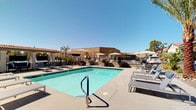This single-level outdoor amenity space is designed for both relaxation and social gatherings, combining thoughtful landscaping with functional, resort-style features. At the heart of the space is a generously sized swimming pool with crystal-clear turquoise water, surrounded by a wide concrete deck lined with modern lounge chairs and market umbrellas that provide ample shade on sunny days. Tall palm trees and manicured greenery frame the pool area, enhancing the warm, sun-soaked atmosphere.

Adjacent to the pool, a covered patio structure with sturdy stone columns and a ceiling fan provides a shaded retreat furnished with multiple sets of contemporary blue cushioned sofas and sleek black-frame coffee tables. This seating area is further extended under a tiled-roof portico, where additional blue lounge furniture is arranged on artificial turf — creating a comfortable, casual setting that blends indoor-style comfort with the outdoors.

A dedicated outdoor kitchen nook houses a full-size stainless steel built-in grill, complete with prep space and utility access, making it well-suited for outdoor cooking and entertaining. Decorative green hedge panels are used throughout the space as privacy screens, adding a polished, finished look. Overall, the space exudes a clean, modern aesthetic with a distinctly warm, desert-resort character.