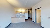This single-floor unit features an open-concept layout that connects the kitchen and living area in a seamless, functional arrangement. The kitchen is equipped with light wood cabinetry, a white refrigerator, electric range, microwave, and a double sink, all positioned along the back wall. A peninsula counter extends outward, creating a natural divide between the kitchen and the carpeted living space while offering additional workspace or casual seating.

The living area is finished with low-pile carpet in a neutral gray tone, contributing to a clean and understated aesthetic. Exposed ductwork is visible near the ceiling in the kitchen zone, lending a subtle industrial character to the space. Track lighting above the kitchen provides focused illumination, while wall-mounted fixtures add ambient light along the hallway leading to the unit's entry door.

The entry is marked by a solid wood door with a dark frame, set along a corridor that connects the main living space to additional areas of the unit. The overall color palette—featuring neutral walls, light cabinetry, and gray flooring—creates a cohesive, low-maintenance environment suited for everyday living.