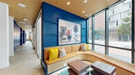 Located at 1509 Broadway in San Diego's dynamic East Village neighborhood, this single-floor amenity space offers a generously sized, thoughtfully designed lounge environment. Floor-to-ceiling glass curtain walls line the street-facing perimeter, flooding the interior with natural light and providing open views of the surrounding urban streetscape, including glimpses of palm trees that nod to the Southern California setting.

The interior design blends bold cobalt blue accent walls with warm wood-paneled ceilings, burnished copper columns, and teal hexagonal tile flooring to create a vibrant yet sophisticated atmosphere. A central lounge zone features a sweeping curved cream sectional sofa anchored by a teal patterned area rug and complemented by a statement sputnik chandelier overhead. Adjacent built-in banquette seating, framed by deep blue millwork and colorful artwork, provides intimate gathering nooks.

A dedicated dining area offers round dark tables with mid-century-style leather chairs, while a glass-enclosed conference room with a bold coastal mural, wall-mounted display, and executive seating serves more formal meeting needs. Private booth-style seating with curtained enclosures adds further flexibility. Throughout, curated artwork, lush potted plants, and layered pendant lighting reinforce a cohesive, design-forward aesthetic that balances social energy with functional versatility.