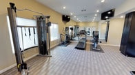 This fitness center offers a comprehensive workout environment designed to support a wide range of exercise routines. The space is finished with warm yellow walls and recessed lighting, creating a bright and energizing atmosphere. Neutral-toned carpet tiles cover the floor throughout, providing a clean, practical surface suited for gym use.

The room is outfitted with a variety of cardio equipment, including treadmills, an elliptical trainer, and a stationary recumbent bike, all arranged to allow comfortable spacing between users. A multi-function cable machine with resistance attachments anchors the strength training area, making the facility suitable for both cardiovascular and resistance-based workouts.

Wall-length mirrors line one side of the room, visually expanding the space while allowing users to monitor their form during exercise. Flatscreen televisions are mounted on the walls, offering entertainment options during workouts. Natural light filters in through windows along one wall, complementing the recessed ceiling lights to keep the space well-lit at all times.

The layout is thoughtfully organized to maximize usable floor area, ensuring ease of movement between equipment. The overall design is clean, functional, and inviting, making this fitness center a practical and well-appointed facility for daily exercise.