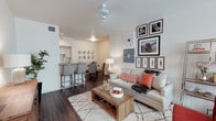 This single-floor residence showcases an open-concept layout that effortlessly connects the main living areas, creating a cohesive and functional flow throughout the space. The living room serves as the centerpiece, featuring warm hardwood-style flooring, a ceiling fan overhead, and neutral wall tones that provide a versatile backdrop for the curated décor. A gallery wall of framed botanical and nature-inspired artwork adds a stylish focal point behind the comfortable seating area, which is anchored by a plush area rug and a rustic wood coffee table. Adjacent to the living room, a dedicated dining area offers ample space for gathering, furnished with upholstered chairs and a well-appointed table. The kitchen, visible from the main living area, features light wood cabinetry and a breakfast bar with seating for casual dining, making it ideal for both everyday living and entertaining. The consistent hardwood-style flooring flows throughout, unifying the open space and enhancing its warm, welcoming ambiance. The overall design strikes a balance between contemporary style and comfortable livability, with carefully selected furnishings and accents that contribute to a refined yet approachable aesthetic.