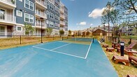 Located at 223 Long Shoals Rd in Asheville, NC, this outdoor recreational facility features a dual-purpose pickleball and tennis court finished in a vibrant blue and green hard-court surface with clearly marked white boundary lines and regulation nets. The court is fully enclosed by a black chain-link fence, providing both safety and definition to the space. Tall light poles are positioned along the perimeter, extending usability into the evening hours. Seating is available courtside, including benches and Adirondack chairs, offering a comfortable vantage point for spectators. Mature trees line portions of the surrounding area, providing natural shade and a lush backdrop that enhances the outdoor ambiance. The facility sits adjacent to a modern multi-story residential building with blue and gray siding, balconies, and large windows, creating a cohesive community atmosphere. The open-air setting allows for ample natural light and fresh air throughout the day, while views of the surrounding Asheville landscape and tree canopy contribute to an inviting and scenic environment. A walkway and additional landscaping connect the court area to the broader community grounds, making this a well-integrated and accessible recreational amenity.