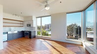 Located on the fourth floor at 60 SE 10th Ave in Portland's vibrant Lower Buckman neighborhood, this modern unit offers an open, light-filled layout enhanced by warm hardwood-style flooring and a clean, contemporary design throughout. The expansive living area is anchored by large floor-to-ceiling corner windows that frame sweeping views of the Portland skyline and surrounding tree canopy, with a ceiling fan adding comfort to the sun-drenched space. The well-appointed kitchen features dark lower cabinetry contrasted by white upper cabinets and open wood shelving, a classic white subway tile backsplash, a farmhouse-style sink, and a full suite of stainless steel appliances including a range, microwave, dishwasher, and refrigerator. The bedroom offers a generous closet with a built-in rod and rail system, and continues the warm flooring found throughout the unit. Two bathrooms serve the space — one featuring a long vanity with ample cabinetry and a large mirror, the other a more compact layout with white cabinetry and a narrow window for natural light. In-unit laundry is conveniently provided via a stacked washer and dryer. The overall ambiance is bright, clean, and contemporary, with an emphasis on natural light and urban views.