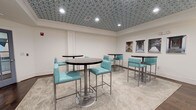 Located at 3112 Maybank Hwy on Johns Island, SC, this business center is a single-floor space designed to blend functionality with contemporary style. The room is immediately distinguished by its bold geometric-patterned ceiling tile, which adds a striking visual element overhead and sets the tone for the space's modern aesthetic.

The lounge area features multiple round, dark-topped high-top tables paired with teal upholstered bar-height chairs with chrome frames, all arranged atop a neutral area rug over hardwood flooring. A series of framed black-and-white pier photography lines the wall, contributing an artistic, coastal-inspired touch that complements the Johns Island location.

The workstation area runs along one wall and is outfitted with a continuous white countertop set over dark cabinetry with teal drawer accents. Multiple desktop computers are available for use, accompanied by a printer/scanner and decorative teal table lamps. A wall-mounted flat-screen TV is positioned above the workstation for shared viewing. A posted "Business Center Rules" sign is clearly visible, indicating this is a shared-use amenity space.

Recessed lighting and flush-mount ceiling fixtures provide consistent illumination throughout. The combination of hardwood flooring, patterned ceiling, teal accents, and modern furnishings creates a cohesive, professionally appointed environment well-suited for productivity and collaboration.