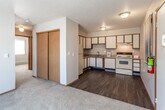 This single-floor unit offers a practical and comfortable layout designed for everyday living. The kitchen is efficiently arranged in an L-shaped configuration, featuring white cabinetry with wood-tone trim, laminate countertops, a double-basin sink, a white electric range, and a full-size refrigerator. Dark hardwood-style flooring runs through the kitchen and entry areas, providing a clean contrast to the lighter walls and cabinetry. The living area transitions seamlessly from the kitchen and is finished with neutral wall-to-wall carpeting, a ceiling fan with light fixture, and a large window fitted with horizontal blinds that allows natural light to filter in. A wall-mounted air conditioning unit adds year-round comfort. The bedrooms are carpeted and feature baseboard heating, individual windows with blinds for natural light and privacy, and a quiet, understated atmosphere. The full bathroom includes a single-sink vanity with under-sink storage, a mirrored medicine cabinet, a bathtub and shower combo with a safety grab bar, and durable hardwood-style flooring. Textured ceilings are consistent throughout the unit. Overall, the space presents a functional and tidy environment with a neutral color palette, making it easy to personalize.