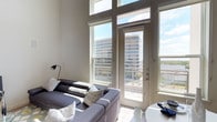 This two-story, 2-bedroom, 2-bathroom residence is designed to maximize light, space, and views. The living area immediately impresses with its dramatically tall ceilings and an expansive wall of oversized windows, flooding the interior with natural light throughout the day. A glass-paned door leads to a private balcony, framing striking views of the surrounding urban skyline and a glimpse of open water beyond — a standout feature of the space.

The bright, airy aesthetic is reinforced by a neutral white palette on the walls and light flooring, creating a clean and modern backdrop that complements the openness of the layout. The living area comfortably accommodates a generous sectional sofa arrangement, with ample room for dining nearby. The two-story configuration adds an additional layer of architectural interest, offering distinct living zones across both levels while maintaining a cohesive, open feel throughout. Natural light and urban panoramas remain consistent themes across the residence, making this a space that is both functional and visually engaging.