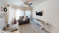 This single-floor residence offers a thoughtfully designed layout that balances comfort and functionality throughout. The living room serves as the heart of the home, featuring plush carpeting, a ceiling fan with light fixture, and large windows dressed with sheer curtains that allow abundant natural light to fill the space. A wall-mounted flat-screen TV and a decorative console table complete the room's welcoming atmosphere.

The kitchen is equipped with warm-toned wood cabinetry, dark granite-style countertops, a double-basin sink, and white appliances including an electric range and built-in microwave. An L-shaped peninsula with bar seating provides a casual dining option and opens to the entryway with hardwood-style flooring. Adjacent to the kitchen, a dedicated dining area features a sliding glass door offering outdoor views, complemented by vertical blinds and a decorative wall mirror.

A carpeted hallway connects the private quarters, which include multiple bedrooms. Each bedroom features ceiling fans, generous natural light from large windows, and soft carpet underfoot. One bedroom benefits from dual windows and ample space for furniture. A built-in desk area in the hallway provides a convenient workspace.

The full bathroom includes a vanity with granite-style countertops, a lighted mirror, a tub-shower combination, and direct access to a spacious walk-in closet, rounding out this well-equipped residence.