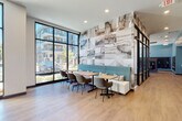 This contemporary fitness center is thoughtfully designed to accommodate a full range of workout activities within a bright, open single-level layout. Large floor-to-ceiling windows with black-framed grid detailing line multiple walls, flooding the space with natural light and providing views of the surrounding outdoor environment. Direct exterior access is available through glass doors, connecting the gym to an outdoor area.

The cardio zone is well-stocked with treadmills, ellipticals, and stationary bikes, all positioned to take advantage of the expansive window views. A dedicated strength training area features a comprehensive dumbbell rack, cable machines, a multi-functional cable cross system, weight benches, and a functional training rig. Additional accessories including resistance bands, stability balls, medicine balls, and a speed bag are neatly organized on wall-mounted racks.

Wall-to-wall mirrors along the strength zone visually expand the space and support proper form during workouts. A bold abstract mural in blue and grey tones adds an artistic focal point. Overhead linear lighting ensures consistent illumination throughout. A water fountain station is also present. The durable, light-toned flooring and clean white walls contribute to a polished, modern aesthetic throughout the facility.