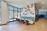 This contemporary fitness center is thoughtfully designed to accommodate a full range of workout activities within a bright, open single-level layout. Large floor-to-ceiling windows with black-framed grid detailing line multiple walls, flooding the space with natural light and providing views of the surrounding outdoor environment. Direct exterior access is available through glass doors, connecting the gym to an outdoor area.

The cardio zone is well-stocked with treadmills, ellipticals, and stationary bikes, all positioned to take advantage of the expansive window views. A dedicated strength training area features a comprehensive dumbbell rack, cable machines, a multi-functional cable cross system, weight benches, and a functional training rig. Additional accessories including resistance bands, stability balls, medicine balls, and a speed bag are neatly organized on wall-mounted racks.

Wall-to-wall mirrors along the strength zone visually expand the space and support proper form during workouts. A bold abstract mural in blue and grey tones adds an artistic focal point. Overhead linear lighting ensures consistent illumination throughout. A water fountain station is also present. The durable, light-toned flooring and clean white walls contribute to a polished, modern aesthetic throughout the facility.