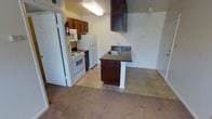 This single-floor space features an open-concept layout that connects the kitchen and living area, creating a cohesive and functional environment. The kitchen is equipped with white appliances, including a electric range, oven, and refrigerator, complemented by warm-toned wooden upper cabinetry for ample storage. A peninsula counter with a dark laminate surface and an integrated sink serves as a natural divider between the kitchen and the adjoining living area, adding both workspace and visual structure to the layout. Dark cabinetry on the peninsula contrasts with white column accents at its base, adding a subtle design detail. A range hood is mounted above, contributing to the kitchen's practical setup. The flooring transitions from tile in the kitchen to carpet in the living area, defining each zone within the open plan. The walls are finished in a neutral tone, lending a clean and understated backdrop throughout. A fire extinguisher is mounted near the entry, reflecting attention to safety. The space offers a straightforward, no-frills layout well-suited for everyday living.