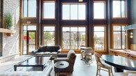 This impressive single-floor community clubhouse showcases a bold blend of rustic and industrial architectural design. The space is anchored by soaring double-height ceilings supported by substantial exposed timber columns, creating a grand, open atmosphere throughout. A dramatic wall of floor-to-ceiling wood-framed windows floods the interior with abundant natural light while offering sweeping views of an outdoor pool deck and surrounding grounds.

The main lounge area features a striking stone fireplace with slate tile surround and a wood mantel, complemented by comfortable leather seating arrangements and layered area rugs. Industrial-style pendant lighting and wall sconces add warmth alongside the exposed brick and natural wood accents that define the aesthetic.

Adjacent to the lounge, a well-appointed kitchen and refreshment area features dark cabinetry with granite countertops, a commercial beverage station, and an exposed brick accent wall adorned with decorative gear-themed artwork — a nod to the space's industrial character.

A dining area with long communal tables and mixed seating provides ample room for gatherings, while large arched mirror panels and rustic reclaimed wood details enhance the overall ambiance. The entryway features glass double doors with clean sightlines through the entire space, reinforcing the open, welcoming layout. Light wood-look flooring ties the zones together seamlessly, creating a cohesive and inviting environment throughout.