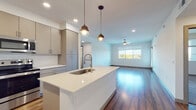 This contemporary single-floor residence showcases a thoughtfully designed open-concept layout that seamlessly connects the kitchen and living areas. The kitchen is a standout feature, equipped with sleek gray flat-front cabinetry, a large center island with a white quartz countertop and undermount sink, stainless steel appliances including a refrigerator, range, and built-in microwave, and a crisp white tile backsplash. Decorative pendant lighting over the island adds a modern, stylish touch. Rich hardwood-style flooring flows throughout the main living areas, catching natural light and adding warmth to the open space. The spacious living room benefits from a ceiling fan, recessed lighting, and access to a window-lined exterior door that brings in ample natural light. Bedroom areas feature plush carpeting and generous walk-in closet space with built-in shelving and hanging rods. Both bathrooms are finished to a high standard, featuring wood-look flooring, gray vanity cabinetry with white countertops, and backlit LED vanity mirrors. One bathroom includes a full tub-shower combination. Throughout the residence, clean white walls, modern hardware, and consistent design choices create a cohesive, contemporary aesthetic.