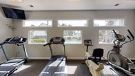 This fitness center is a dedicated, single-floor exercise facility designed to support a well-rounded workout routine. The space is outfitted with a comprehensive selection of equipment, including treadmills, a recumbent bike, an elliptical trainer, a rowing machine, and a multi-station cable strength machine, catering to both cardiovascular and resistance training needs. Protective rubber matting is placed beneath equipment, complementing the carpeted flooring throughout the room. A large wall-mounted mirror enhances the sense of space while allowing users to monitor their form during workouts. Natural light pours in generously through a combination of wide horizontal clerestory windows positioned near the ceiling and larger standard windows along the walls, offering views of the surrounding outdoor landscape, including a pool area framed by palm trees and greenery. Overhead fluorescent panel lighting supplements the natural illumination. A beverage vending machine stocked with water and sports drinks is conveniently located near the entrance, providing easy access to hydration. Posted fitness center rules near the door help maintain a welcoming and orderly environment. The overall ambiance is bright, clean, and functional, creating an inviting space for residents to prioritize their health and wellness.