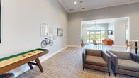 This well-appointed single-floor community clubhouse in Noblesville, Indiana offers a generously sized, open layout designed for both socializing and relaxation. The space is unified by light wood-look flooring, soft neutral wall tones, and crown molding throughout, creating a cohesive and inviting atmosphere.

The main lounge area serves as a focal point, anchored by a dramatic floor-to-ceiling dark tile fireplace surround that draws the eye upward, emphasizing the soaring ceiling height. Vibrant artwork adorns the walls, adding energy and color to the space. Adjacent to the lounge is a dedicated game area equipped with a shuffleboard table, pool table, and a wall-mounted flat-screen TV, perfect for recreation and entertainment.

A fully equipped kitchen features gray shaker-style cabinetry, stainless steel appliances, a subway tile backsplash, and a large island with a white countertop and textured wood panel siding, complemented by rattan bar stools and stylish pendant lighting. A casual dining zone with bistro-height tables and woven chairs sits nearby, bathed in natural light from large arched transom windows and French doors that frame expansive outdoor views.

A private office with glass sliding doors provides a quiet workspace. Throughout, the space balances modern design with warm, comfortable furnishings, making it a versatile and welcoming community destination.