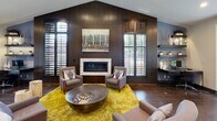 Located at 2800 S Syracuse Way in Denver, Colorado, this single-floor space presents a thoughtfully designed interior that blends contemporary style with functional versatility. The lounge area serves as a focal point, anchored by a sleek linear fireplace set against rich, dark wood-paneled walls. Plantation shutters frame multiple windows, allowing natural light to filter in while offering views of the surrounding greenery. A vibrant yellow area rug and a sculptural metal coffee table add bold contrast to the warm, earthy tones throughout. Adjacent to the lounge, a dedicated workspace features floating wood shelves, a glass-top desk, and accent shelving displaying decorative vessels, all set against a cool blue accent wall. The kitchen area is outfitted with slate-toned cabinetry, stacked stone tile accents, a full-size stainless steel refrigerator, and warm wood-finished ceiling details, giving it a modern yet inviting character. A dining nook bathed in natural light through large grid-pane windows is furnished with a rustic round wood table and upholstered chairs, framed by flowing ombre drapery. Dark tile flooring with a wood-grain appearance runs consistently throughout, unifying the space with a cohesive, polished aesthetic.