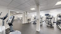 Located at 151 S Bishop Ave in Secane, PA, this fitness center offers a generously sized, single-floor facility designed to accommodate a full range of workout activities. The space is organized across an open, interconnected layout with distinct zones for cardio training, strength machines, and free weights, allowing users to move efficiently between equipment areas.

The cardio section is well-equipped with multiple treadmills and elliptical trainers from TRUE Fitness, arranged in orderly rows to maximize usability. A dedicated strength training area features a cable/functional trainer machine alongside a variety of selectorized machines targeting major muscle groups, while a separate free weight zone includes a dumbbell rack and an adjustable bench. Wall-mounted mirrors in select areas support proper form and enhance the sense of space.

The facility features durable rubber flooring throughout, providing comfort and safety underfoot. Overhead, a drop-panel ceiling with recessed fluorescent lighting ensures consistent, even illumination across the entire floor. White walls contribute to a clean, bright, and neutral environment. A wall-mounted television is available in the free weight area for added convenience. The layout is punctuated by structural columns that subtly define workout zones without interrupting the overall openness of the space. The fitness center presents a practical, well-organized environment suited for a comprehensive workout routine.