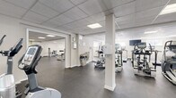 Located at 151 S Bishop Ave in Secane, PA, this fitness center offers a generously sized, single-floor facility designed to accommodate a full range of workout activities. The space is organized across an open, interconnected layout with distinct zones for cardio training, strength machines, and free weights, allowing users to move efficiently between equipment areas.

The cardio section is well-equipped with multiple treadmills and elliptical trainers from TRUE Fitness, arranged in orderly rows to maximize usability. A dedicated strength training area features a cable/functional trainer machine alongside a variety of selectorized machines targeting major muscle groups, while a separate free weight zone includes a dumbbell rack and an adjustable bench. Wall-mounted mirrors in select areas support proper form and enhance the sense of space.

The facility features durable rubber flooring throughout, providing comfort and safety underfoot. Overhead, a drop-panel ceiling with recessed fluorescent lighting ensures consistent, even illumination across the entire floor. White walls contribute to a clean, bright, and neutral environment. A wall-mounted television is available in the free weight area for added convenience. The layout is punctuated by structural columns that subtly define workout zones without interrupting the overall openness of the space. The fitness center presents a practical, well-organized environment suited for a comprehensive workout routine.