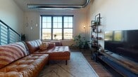 This two-floor urban loft showcases a striking industrial-modern aesthetic defined by soaring vaulted ceilings, exposed ductwork, and an open-concept layout that seamlessly connects the kitchen, dining, and living areas on the main level. A bold open-riser steel staircase with horizontal rail balusters serves as a dramatic architectural centerpiece, leading to the upper loft level. The main living area features rich hardwood flooring, large multi-pane windows that frame outdoor views and flood the space with natural light, and a ceiling fan overhead. Pendant jar lights hang above a butcher-block kitchen island with bar seating, complementing the well-appointed kitchen outfitted with gray shaker cabinetry, open wood shelving, white countertops, and stainless steel appliances. Two bathrooms echo the space's design language with gray vanities, white countertops, and wood-look flooring. The upper loft-level bedroom benefits from plush carpeting, skylights that bathe the room in daylight, a ceiling fan, and an open railing that offers a view down to the main floor. A second carpeted bedroom on the upper level also features vaulted ceilings and exposed ductwork, adding to the loft's cohesive industrial character throughout.