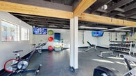 Located at 3202 W Bell Rd in Phoenix, Arizona, this fitness center offers a well-equipped, single-floor workout space designed to accommodate a wide range of exercise routines. The room features an exposed industrial ceiling with dark steel beams and structural elements accented by warm natural wood beams, giving the space a modern, utilitarian aesthetic. Bright linear LED lighting runs throughout, ensuring the entire floor is evenly illuminated.

The flooring consists of durable dark rubber gym matting, ideal for both heavy equipment and bodyweight training. The space is divided into distinct functional zones: a cardio area equipped with treadmills, elliptical machines, and stationary spin bikes; a strength training section featuring a dumbbell rack with a full range of free weights and adjustable benches; and a functional training area with TRX suspension trainers, stability balls, medicine balls, and a wall-mounted plyometric board. Motivational phrases such as "Flexibility" and "Core Stability" are painted directly onto the white cinder block walls in bold yellow lettering, reinforcing the space's purpose-driven design.

Multiple wall-mounted flat-screen TVs are positioned throughout the room, keeping members engaged during workouts. Windows along the perimeter allow natural light to filter in, contributing to an energetic and open atmosphere well-suited for a comprehensive fitness experience.