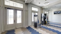 This fitness center offers a thoughtfully designed workout environment that balances functionality with aesthetic appeal. The space is flooded with natural light, courtesy of large transom windows and glass-paneled entry doors that frame views of the surrounding exterior. Soaring ceilings add a sense of openness and airiness throughout the facility.

The flooring makes a striking visual statement, featuring a chevron-patterned design that blends neutral wood tones with bold blue accents, creating a dynamic and energetic atmosphere underfoot. The walls are finished in a clean, neutral gray palette that keeps the space feeling fresh and modern.

Strength training is well-represented, with a cable machine and multi-function weight equipment visible throughout. A large wall-mounted mirror expands the sense of space and allows members to monitor their form during workouts. Recessed lighting and a modern circular pendant fixture provide ample illumination to complement the natural light.

The entry area is clean and welcoming, with classic architectural detailing including crown molding and trimmed doorframes that give the fitness center an upscale feel. Overall, this facility delivers a polished and motivating environment well-suited for a comprehensive workout routine.