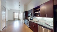 This well-appointed single-floor residence showcases a contemporary design with impressively tall ceilings that lend an airy, open atmosphere throughout the living areas. Polished concrete floors run through the main living space and kitchen, providing a clean, modern aesthetic that is both durable and visually striking.

The kitchen is a standout feature, outfitted with rich dark espresso cabinetry — including full-height upper cabinets and open display shelving — paired with granite countertops and a classic white subway tile backsplash. A full suite of stainless steel appliances, including a range, built-in microwave, dishwasher, and refrigerator, completes the culinary space. Track lighting overhead highlights the kitchen's sleek finishes.

The living area benefits from abundant natural light streaming through large windows dressed with curtain panels, creating a bright and welcoming environment. The bedroom features plush carpeting, soaring ceilings, and generous window exposure, adding warmth and comfort to the private quarters. A spacious walk-in closet with wire shelving provides ample storage.

The bathroom is finished with matching granite countertops, dark cabinetry with brushed nickel hardware, tile flooring, and elegant wall sconces. A dedicated laundry area with wire shelving and utility connections adds everyday convenience to this thoughtfully designed residence.