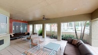 This single-floor space centers around a generously sized covered outdoor lounge area designed for comfort and relaxed social gatherings. The structure features a textured ceiling equipped with ceiling fans to ensure comfortable airflow, making the space enjoyable in warm climates. Sturdy square columns line the perimeter, framing open views of the surrounding landscaped grounds, which include mature trees, manicured shrubs, and a white fence that adds a sense of enclosure and privacy. The flooring consists of light-toned ceramic tiles that lend a clean, durable surface well-suited for outdoor use. The seating arrangement is thoughtfully organized, with a combination of cushioned armchairs in striped fabric and deep brown upholstered sofas, all paired with low-profile square coffee tables. The open-air design allows abundant natural light to filter through from multiple sides, creating a bright and inviting ambiance throughout the day. Colorful accent walls in warm red and green tones add a vibrant visual character to the architecture. Overall, the space strikes a balance between functional outdoor design and relaxed, resort-style comfort, making it well-suited as a communal lounge or gathering area.