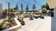 Located at 2424 N Tustin Ave in Santa Ana, California, this thoughtfully designed outdoor amenity space offers a resort-inspired atmosphere ideal for relaxation and socializing. At its centerpiece is a generously sized swimming pool with clear, turquoise water, surrounded by a spacious concrete sun deck furnished with lounge chairs and vibrant yellow umbrellas that provide ample shade on sunny Southern California days. Tall mature trees frame the perimeter, lending a sense of privacy and a natural canopy overhead. Adjacent to the pool, a dedicated outdoor lounge and dining area features comfortable seating arrangements accented by string lights overhead, creating an inviting atmosphere that transitions seamlessly from day to evening. Bold pops of color — from orange chaise lounges to the building's accented exterior facades in orange, dark navy, and white — give the space a modern, lively character. Thoughtfully placed planters with tropical and drought-tolerant plants add greenery throughout. A private patio area enclosed by sleek glass and dark metal railings offers a quieter retreat with views of the landscaped surroundings. The overall ambiance is bright, open, and contemporary, reflecting the sunny Southern California lifestyle.