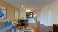 This single-floor residence offers an open-concept design that seamlessly connects the living, dining, and kitchen areas into one cohesive and functional living space. The warm, golden-toned walls complement rich hardwood flooring that flows throughout, creating an inviting and unified ambiance.

The kitchen is a standout feature, outfitted with dark espresso cabinetry, stainless steel appliances including a refrigerator and range, and a kitchen island with pendant lighting overhead. The island provides additional prep and seating space, while a skylight above the kitchen enhances the area with natural light.

Adjacent to the kitchen, the dining area accommodates a round table with seating for four, lit by track lighting above. Decorative accents add a personal touch to the space. The living area, visible in the foreground, features a glass-top coffee table and comfortable seating, with a framed mirror on the wall that enhances the sense of openness.

A hallway leads to additional rooms, suggesting a practical and well-organized floor plan. White interior doors and trim provide clean contrast against the warm wall tones throughout. Overall, the space balances modern finishes with a cozy, livable atmosphere.