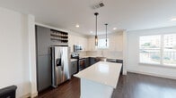Located at 322 E Broadway Ave in Fort Worth, TX, this modern two-story residence showcases contemporary design throughout its well-planned layout. The open-concept kitchen is a standout feature, boasting a two-tone cabinet scheme with crisp white upper cabinets and slate gray lower cabinets, complemented by a classic white subway tile backsplash. A large center island with a sleek white quartz countertop provides ample workspace and serves as a natural gathering point. Stainless steel appliances, including a French door refrigerator, range, microwave, and dishwasher, round out the kitchen's functionality. A built-in wine rack adds a refined touch above the refrigerator. Dark hardwood-style flooring runs throughout the main living areas, creating a rich contrast against the bright white walls. The living spaces are open and airy, with natural light filtering in through multiple windows. Bedrooms feature plush carpeting and ceiling fans, with windows offering outside views. The bathrooms are finished with gray vanities, white quartz countertops, large framed mirrors, and full white subway tile surrounds in the tub and shower areas. Light tile flooring in the bathrooms ties the clean, modern aesthetic together seamlessly across both floors.