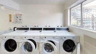 This shared laundry room is a well-organized, single-floor facility designed to accommodate multiple users efficiently. The space features light-colored tile flooring throughout, contributing to a clean and practical environment. Fluorescent lighting illuminates the room evenly, ensuring good visibility across all areas.

The main laundry area is equipped with a row of front-loading washing machines along one wall, each featuring digital controls for convenient cycle selection. Opposite the washers, a bank of stacked dryers provides ample drying capacity. The machines are coin-operated, making the facility accessible for shared use.

A deep utility sink is positioned near one end of the room, offering a dedicated space for hand-washing or pre-treating garments. A separate folding area includes a large, white worktable with a metal frame, providing a generous surface for sorting and folding laundry.

Natural light enters through windows on one side of the room, brightening the space and offering views of the surrounding exterior. Two entry doors—one white-paneled and one black—provide convenient access points. The white walls and minimal design keep the overall ambiance clean, straightforward, and functional, prioritizing utility and ease of use throughout.