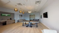 Central Gardens Grand Café is a thoughtfully designed single-floor amenity lounge that blends recreation, relaxation, and social gathering into one cohesive space. The open floor plan is anchored by light hardwood flooring throughout, creating a warm and inviting atmosphere beneath a sleek drop ceiling with recessed lighting.

The game area takes center stage with a full-size ping pong table positioned beneath a striking cluster of globe pendant lights in brushed gold, adding a bold, artistic touch to the space. Nearby, a dedicated dining and meeting area features a dark console table with upholstered chairs and wall-mounted flat-screen televisions, suitable for casual gatherings or small group meetings.

The lounge zone is defined by a contemporary neutral sofa, a geometric patterned area rug, and a sculptural metallic coffee table. Bold pop-art canvases and vibrant wall-mounted artwork contribute to the space's energetic yet refined character.

A built-in café bar — branded as the Central Gardens Grand Café — offers a compact kitchenette with a beverage refrigerator, coffee station, and wood cabinetry, perfect for casual refreshments.

Floor-to-ceiling windows and wide sliding glass doors open to an outdoor terrace with seating, allowing abundant natural light and scenic treetop views to enhance the overall ambiance of the interior.