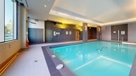 This generously sized indoor aquatic facility in Canada offers a well-appointed and thoughtfully designed space for swimming and relaxation. The centerpiece is a large rectangular lap pool with crystal-clear water, bordered by wide tiled pool decking in neutral tones. Alongside the lap pool sits a separate hot tub or spa pool, framed by floor-to-ceiling windows that flood the space with natural light and offer views of the surrounding greenery and streetscape. Tall tropical plants are positioned near the windows, adding a vibrant, resort-like ambiance to the interior. The architecture features bold dark-tiled columns that provide strong visual contrast against the lighter walls and flooring, lending the space a modern and sophisticated character. Recessed and layered ceiling lighting, including warm accent lighting, enhances the atmosphere throughout. A dedicated lounge area with contemporary chaise lounges and benches provides a comfortable space to relax. The facility also includes change room access, emergency safety equipment, and clearly marked signage. The overall design balances functionality with an upscale aesthetic, creating an inviting environment suited for both fitness and leisure.