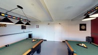 The billiards room at 6304 Rancho Mission in San Diego is a dedicated recreational space designed for pool enthusiasts. The room accommodates at least two full-sized pool tables with green felt surfaces and wooden rail trim, providing ample space for gameplay and movement around each table. Overhead, a classic hanging billiard light fixture with black shades illuminates one of the tables, while recessed ceiling speakers suggest an integrated audio setup for an enhanced atmosphere. The textured white ceiling features exposed beam detailing, adding subtle architectural character to the space. Neutral white walls are accented with small decorative pieces, keeping the focus on the recreational function of the room. The floor is finished in a durable speckled gray surface well-suited to high-traffic use. A small bar or counter area is visible along one wall, accompanied by a compact refrigerator, making the space well-equipped for entertaining. A set of double doors provides access, and in-ceiling speakers are positioned throughout, completing this well-appointed game room.