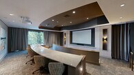Located at 80 Mill St in Hanover, Massachusetts, this thoughtfully designed private screening room delivers an upscale cinematic experience within a refined, contemporary setting. The space is anchored by a large projection screen mounted at the front wall, framed by warm wood paneling and decorative wall sconces that add a soft, ambient glow. Seating is arranged in a curved, tiered configuration, with rich cognac-colored leather armchairs providing both comfort and a luxurious aesthetic. A curved partition with a marble-topped surface divides the seating tiers, adding an architectural focal point. The ceiling is a standout design element, featuring a dramatic layered tray design with recessed lighting set against a deep charcoal finish, lending depth and a cinematic atmosphere to the room. Plush, textured carpet in a muted gray tone runs throughout, enhancing acoustics while complementing the overall color palette. Floor-to-ceiling windows with elegant gray drape curtains line one wall, offering views of lush surrounding greenery and filling the space with natural light when desired. A rear lounge area provides additional seating with leather armchairs arranged around a small table, accompanied by framed artwork on the walls. This screening room combines modern architecture with premium finishes for an immersive and sophisticated entertainment environment.