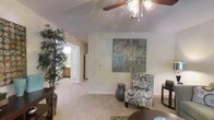 This single-floor residence offers a welcoming and well-appointed living space designed for comfortable everyday living. The main living area is highlighted by vaulted ceilings that lend a sense of openness and airiness, complemented by a ceiling fan with a light fixture that provides both functionality and a decorative touch. Soft wall-to-wall carpeting runs throughout, adding warmth and comfort underfoot. The neutral, light-toned walls create a bright and versatile backdrop, ideal for a variety of interior styles. A large piece of abstract wall art serves as a focal point, adding a contemporary artistic element to the space. The living area flows naturally into the kitchen, which is visible through an open passageway and features wood-toned cabinetry and standard appliances, including a refrigerator. A tall indoor plant positioned near the entryway introduces a refreshing natural element, enhancing the overall ambiance. The layout is thoughtfully arranged to maximize usability, with clear sightlines connecting the living and kitchen areas. The textured ceiling adds a subtle architectural detail that completes the interior character of this inviting single-story home.