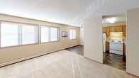 This single-floor unit offers a practical and well-organized layout designed for comfortable everyday living. The open living area is generously sized, featuring neutral beige walls, soft wall-to-wall carpeting, and a textured ceiling. Multiple windows along one wall allow natural light to fill the space, while a wall-mounted air conditioning unit and baseboard heating provide year-round climate control. A ceiling fan is positioned at the transition point between the living area and the kitchen.

The kitchen is efficiently arranged with warm oak cabinetry spanning both upper and lower sections, a double-basin sink, a full-size refrigerator, dishwasher, and a tile floor in a dark stone pattern. Fluorescent overhead lighting ensures the kitchen is well-lit.

The bedroom is carpeted and includes a window with natural light and a closet with wood-panel doors. A hallway connects the bedroom to additional storage and the full bathroom, which features a single-sink vanity with oak cabinet storage, a wall mirror with bar lighting, a soaker tub with grab bars, and tile flooring. The entry area includes a closet and transitions smoothly into the main living space.