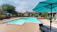 This outdoor amenity space in Frisco, Texas, centers around a generously sized swimming pool with clear, turquoise water, offering a resort-like atmosphere for residents. The pool area is defined by classic brick coping and a textured aggregate deck that surrounds the water on all sides, providing a durable and attractive surface for lounging and foot traffic. A built-in spa with a decorative brick surround sits adjacent to the main pool, accompanied by a fountain feature that adds a soothing water element to the setting. Poolside comfort is well-considered, with rows of chaise lounge chairs positioned along the deck and shaded by vibrant teal market umbrellas. An additional covered seating area is visible nearby, offering a shaded retreat for those seeking respite from the sun. The space is enclosed by iron fencing and lush landscaping, including manicured shrubs, ornamental trees, and established shade trees that provide a natural, park-like backdrop. The clubhouse building, featuring a prominent pitched roofline, large windows, and a covered porch, overlooks the pool and adds an architectural anchor to the overall outdoor environment. The surrounding multi-story residential buildings, clad in neutral tones, frame the space and reinforce the cohesive community setting.