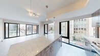 This contemporary 2-bedroom, 2-bathroom residence offers a thoughtfully designed single-floor layout that maximizes both comfort and style. The open-concept kitchen and living area serves as the heart of the home, anchored by a generous island topped with striking marble-veined quartz countertops and complemented by warm wood cabinetry, a white tile backsplash, stainless steel appliances, and elegant pendant lighting with brass accents. Floor-to-ceiling black-framed windows and sliding glass doors flood the living space with natural light while framing expansive urban views of the surrounding cityscape. Light wood-style flooring runs throughout the main living areas, lending a clean, modern aesthetic. A well-lit hallway connects the bedrooms and bathrooms, featuring recessed lighting and light wood flooring that carries a bright, airy feel. The primary bedroom is generously proportioned, with large sliding glass doors opening to outdoor views and plush carpeting underfoot. The second bedroom is finished with the same soft carpeting and neutral tones. Both bathrooms are elegantly appointed — one featuring a glass-enclosed walk-in shower with large-format tile, and the other a soaking tub with floor-to-ceiling marble-look tile surround. The overall ambiance is clean, bright, and urban-modern throughout.