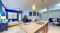 This single-floor residence showcases a cohesive, vibrant design defined by striking cobalt blue accent walls that flow throughout the living room, dining area, kitchen, and bedroom. The open-concept layout connects the kitchen, dining, and living spaces seamlessly, creating an inviting, functional environment.

The kitchen is well-appointed with rich cherry wood cabinetry, granite countertops, stainless steel appliances including a range, microwave, and refrigerator, and a breakfast bar illuminated by yellow pendant lights. A track lighting system above the kitchen adds additional ambiance. Large format tile flooring runs throughout the main living areas.

The living room features vaulted ceilings with a ceiling fan, bold blue accent walls, and access to an exterior door. The adjacent dining nook benefits from a skylight overhead, with a round glass-top table surrounded by elegant nailhead-trim upholstered chairs.

The bedroom offers plush carpeting, a bold blue feature wall, and a walk-in closet. The bathroom includes a tub-shower combination, dark wood vanity cabinetry, and tile flooring. Throughout, the design maintains a bold, modern coastal aesthetic.