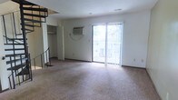 This two-story space offers a functional and straightforward layout designed for comfortable living. The main level features a generously sized living area finished with wall-to-wall carpeting, providing a warm and soft underfoot feel throughout the space. A sliding glass door along the back wall floods the room with natural light and offers a view of an outdoor area, likely a balcony or patio, framed by surrounding greenery visible through the glass. Vertical blinds accompany the door, allowing for flexible light control. A wall-mounted air conditioning unit sits above the door, ensuring climate comfort during warmer months. Adjacent to the living area, a closet with built-in shelving provides convenient storage space. A staircase with metal railing connects the main level to the upper floor, adding vertical dimension to the layout. The white walls throughout keep the space feeling bright and neutral, offering a clean canvas adaptable to various interior styles. The upper level expands the overall living footprint, suitable for use as sleeping quarters or additional living space. Overall, the space presents a simple, practical design with key amenities in place.
