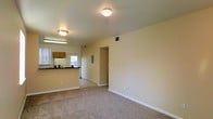 This two-floor residence offers a practical and well-organized layout designed for comfortable everyday living. The main level features an open living area finished with wall-to-wall carpeting, creating a warm and inviting atmosphere. Neutral-toned walls and flush-mount ceiling lighting provide a bright, clean aesthetic throughout. The living space flows seamlessly into the kitchen area, which is separated by a breakfast bar counter — ideal for casual dining or meal prep. The kitchen is equipped with overhead wood-tone cabinetry, a utility sink, and a window that allows natural light to filter in. An electrical panel is conveniently accessible near the kitchen transition area. Ceiling-mounted ventilation registers are present throughout, indicating a central HVAC system. The open doorway from the main living area leads further into the floor plan, suggesting additional rooms and functional spaces beyond. With its two-floor configuration, the residence provides generous separation between living and private areas. The overall design is straightforward and functional, with clean lines, consistent finishes, and a neutral color palette that offers a versatile canvas for personalization.