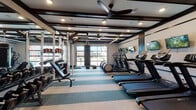 This fitness center is thoughtfully designed to support a wide range of workout routines within a single, open floor plan. The space is anchored by a coffered ceiling with dark wood beam detailing and recessed lighting, complemented by a large ceiling fan that promotes air circulation throughout. A standout architectural feature is the full-width glass panel garage-style door along one wall, which floods the interior with natural light and provides views of the surrounding exterior.

The cardio zone lines one side of the room and includes multiple treadmills and elliptical machines, each paired with wall-mounted flat-screen televisions for entertainment during workouts. On the opposite side, a well-stocked free weight area features a rack system with dumbbells, kettlebells, medicine balls, and a weight bench, alongside a power rack for strength training. A heavy punching bag hangs centrally from a dedicated ceiling mount, adding a functional boxing element to the space.

The flooring consists of striped, multi-tone carpet tiles that visually organize the workout zones. A separate enclosed studio room is accessible through full-height glass sliding doors with dark metal frames, featuring wood flooring, warm wall sconces, and framed wall art, offering a quieter setting for stretching or low-impact training.