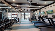 This fitness center is thoughtfully designed to support a wide range of workout routines within a single, open floor plan. The space is anchored by a coffered ceiling with dark wood beam detailing and recessed lighting, complemented by a large ceiling fan that promotes air circulation throughout. A standout architectural feature is the full-width glass panel garage-style door along one wall, which floods the interior with natural light and provides views of the surrounding exterior.

The cardio zone lines one side of the room and includes multiple treadmills and elliptical machines, each paired with wall-mounted flat-screen televisions for entertainment during workouts. On the opposite side, a well-stocked free weight area features a rack system with dumbbells, kettlebells, medicine balls, and a weight bench, alongside a power rack for strength training. A heavy punching bag hangs centrally from a dedicated ceiling mount, adding a functional boxing element to the space.

The flooring consists of striped, multi-tone carpet tiles that visually organize the workout zones. A separate enclosed studio room is accessible through full-height glass sliding doors with dark metal frames, featuring wood flooring, warm wall sconces, and framed wall art, offering a quieter setting for stretching or low-impact training.