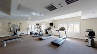This fitness center is a comprehensively equipped exercise facility designed to support a full range of workout routines. The space is finished with neutral, cream-colored walls and low-profile carpeted flooring throughout, creating a clean and functional environment. Natural light filters in through multiple windows positioned along the walls, complementing the recessed ceiling lighting and overhead HVAC vents that maintain a comfortable atmosphere during workouts.

The cardio section is well-appointed with multiple treadmills, elliptical cross-trainers, and a stationary bike, several of which feature integrated display consoles. Wall-mounted flat-screen televisions are positioned throughout the space for entertainment during cardio sessions. A water cooler is conveniently available for hydration.

The strength training area includes a cable resistance machine, a power rack, an adjustable weight bench, and a medicine ball. A dedicated free weights station offers a full rack of dumbbells in varying sizes, along with exercise balls in multiple sizes for functional fitness training.

A large floor-to-ceiling mirror spans one wall, effectively doubling the perceived sense of space and allowing users to monitor their form. Standing fans supplement the climate control system. The layout is open and thoughtfully organized, providing ample room to move between equipment comfortably.