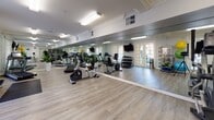 Located at 2290 Mackenzie Creek Rd in Chula Vista, CA, this fitness center is a thoughtfully designed, single-floor facility that offers a comprehensive workout environment. The space features light wood-style flooring throughout, crown molding details, and crisp white walls that give the room a clean, polished aesthetic.

Floor-to-ceiling mirrors line the walls, amplifying the sense of space and allowing users to monitor their form during exercise. The cardio section is well-appointed with multiple treadmills, recumbent bikes, and an elliptical trainer from quality brands. A wall-mounted television provides entertainment during workouts.

A dedicated dumbbell rack offers a range of free weights, while a colorful station houses medicine balls, stability balls, resistance bands, and suspension training equipment — catering to a variety of functional fitness routines. Built-in open shelving provides convenient storage for personal items or accessories.

Natural light filters in through double French doors, connecting the interior to the outdoors and enhancing the bright, airy atmosphere. Overhead fluorescent lighting ensures the space remains well-lit throughout the day. Potted indoor plants are strategically placed throughout, adding warmth and a touch of nature to the environment. Overall, the fitness center strikes a balance between functionality and an inviting, well-maintained ambiance.