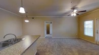 This single-floor space showcases an open-concept layout that seamlessly connects the kitchen and living areas. The kitchen features granite countertops with a peninsula-style counter that extends into the main living space, providing both prep space and a casual dining or seating area. Pendant lighting hangs above the counter, adding a focused light source that complements the overall illumination of the space.

The living area is finished with wall-to-wall carpeting in a neutral tone, contributing to a warm and comfortable atmosphere. Crown molding lines the ceiling throughout, lending a polished, finished look to the interior. A ceiling fan with a light fixture is centrally positioned in the living area, offering both air circulation and supplemental lighting.

The walls are painted in a soft, warm beige tone that enhances the sense of openness. A doorway visible from the living area leads to an adjacent room, suggesting a well-connected and functional floor plan. The space benefits from recessed lighting in the ceiling, ensuring consistent illumination throughout. Overall, the interior presents a clean, move-in-ready environment with thoughtful finishing details.